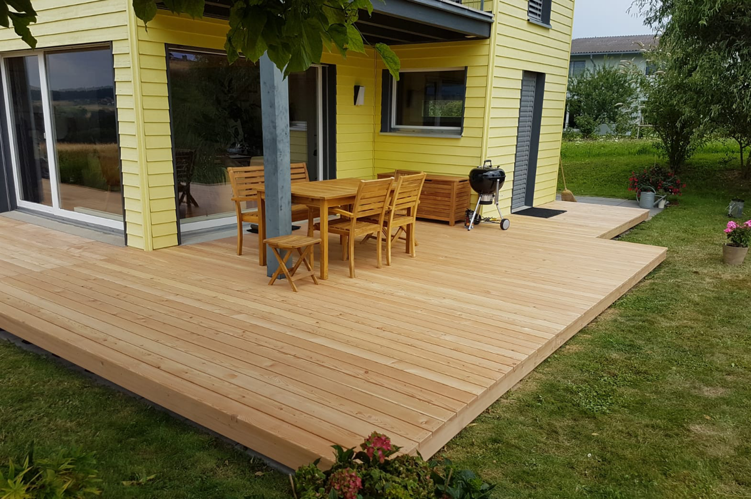 1 Holzterrasse mit Gartenmöbeln und Grill neben einem gelben Haus, umgeben von grünem Garten. Ideal für entspannte Abende.